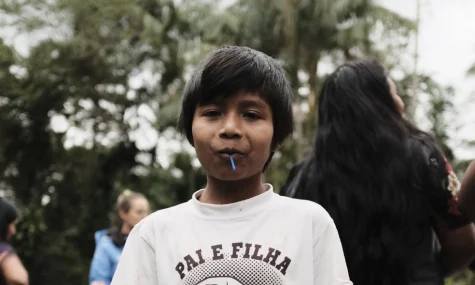 Menino indígena de cabelo escuro vestindo camiseta branca com a frase "Pai e Filha", segurando uma pequena flor azul na boca, ao ar livre com árvores ao fundo