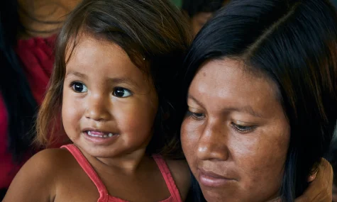 Menina pequena no colo de uma mulher, ambas com traços indígenas, em close mostrando os rostos próximos