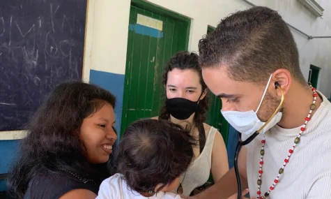 Profissional de saúde com máscara e estetoscópio examinando uma criança no colo da mãe, dentro de uma sala com lousa e porta verde ao fundo