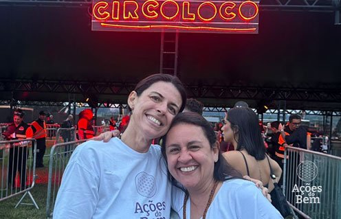 Duas mulheres do Ações do Bem sorrindo abraçadas em frente ao letreiro luminoso do Circoloco