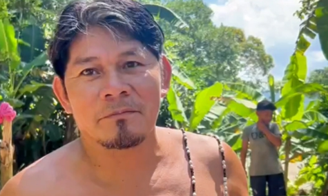 Homem de cabelos escuros e curtos, sem camisa, olhando diretamente para a câmera em um ambiente ao ar livre. Ao fundo, há vegetação tropical com grandes folhas verdes e outra pessoa em pé mais distante. A cena é iluminada pela luz do dia, com céu parcialmente nublado visível entre as árvores.
